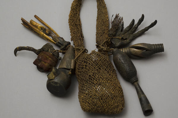 A bag made of vegetable fibers containing a wrapped bundle, with charms of wood, feathers, and animal feet attached