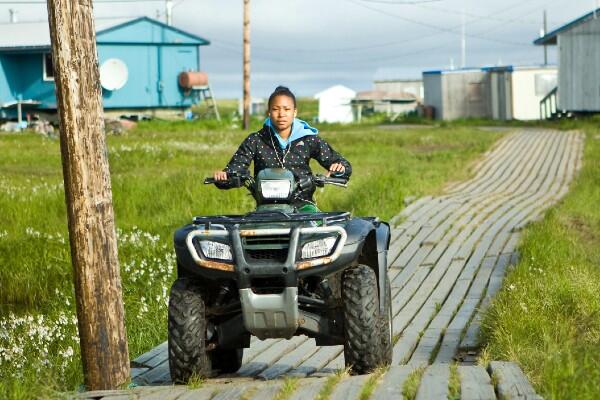 Remote coastal Alaskan Native Village of Newtok and a teen driving a four-wheeler along wooden path.