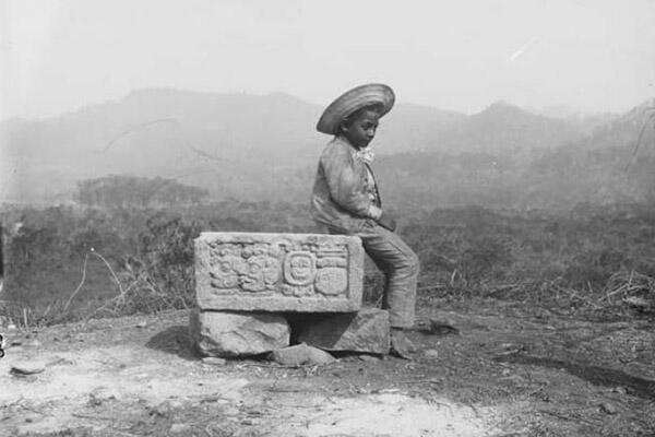 boy in sombrero sits on maya sculpture in historic photo.