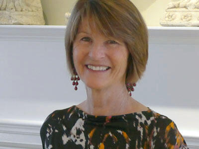 A photo of Anne Best smiling at the camera. She wears beaded red dangly earrings and a patterned shirt, her caramel colored hair swooping across her forehead.