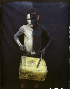 A boy stands playing a yellow drum. Standing against a black background, his body is painted black and white, split vertically.