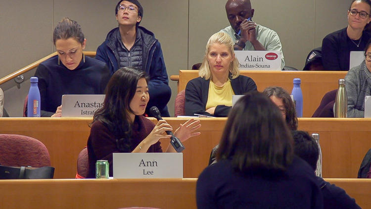 Medium close up of student with name card "Ann Lee" speaking into microphone. Other students watch and listen. Back of Professor Battilana's head is visible in foreground.