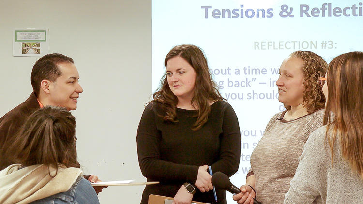 Four graduate students and Instructor Brion-Meisels standing together in a small group in discussion. There is a section of a projected slide visible behind them that reads "Tension & Reflections."