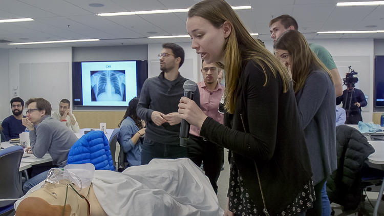 Close up of graduate student with a microphone leaning over and speaking to a fake patient on a hospital bed. Other graduate students in the classroom look on.