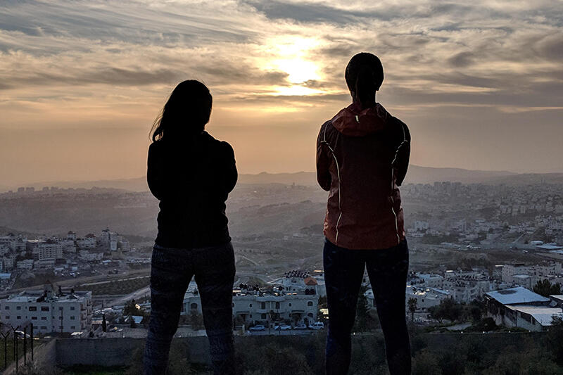 Two students looking out at a city landscape, silhouetted by the sunset