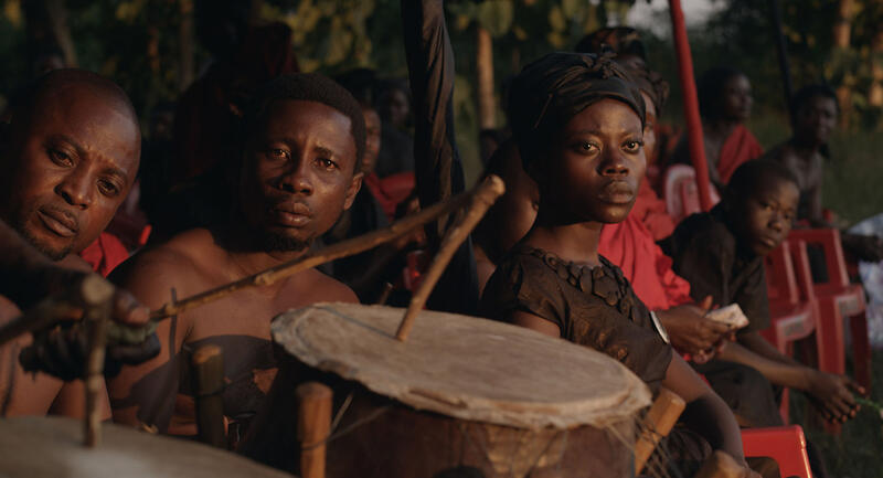A small group of people face the camera, drums being played in the foreground