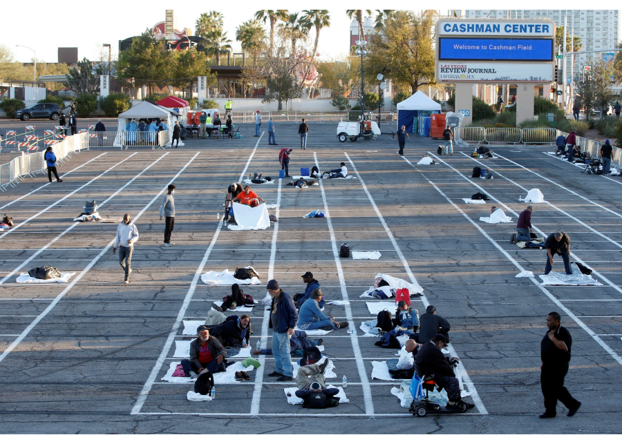 Photo of insecurely housed people sleeping in designated spots in a sunny parking lot with a sign that reads “Cashman Center.”