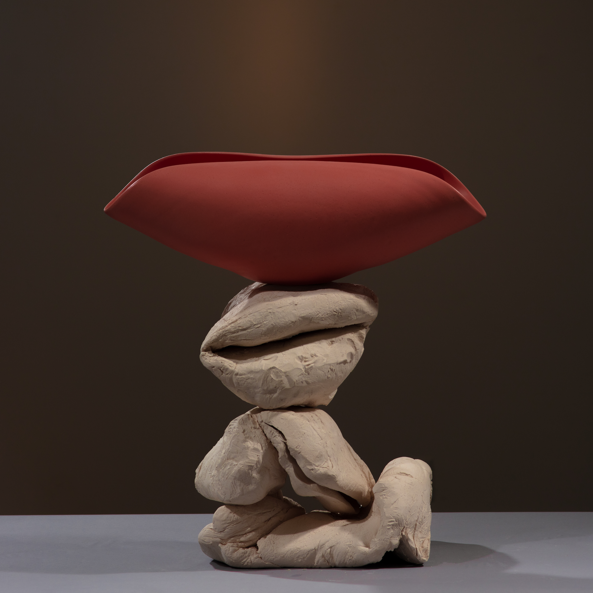 stacked hunks of unfired kneaded clay with a matte red glazed folded bowl shaped vessel balanced on top, arranged on a black fabric backdrop