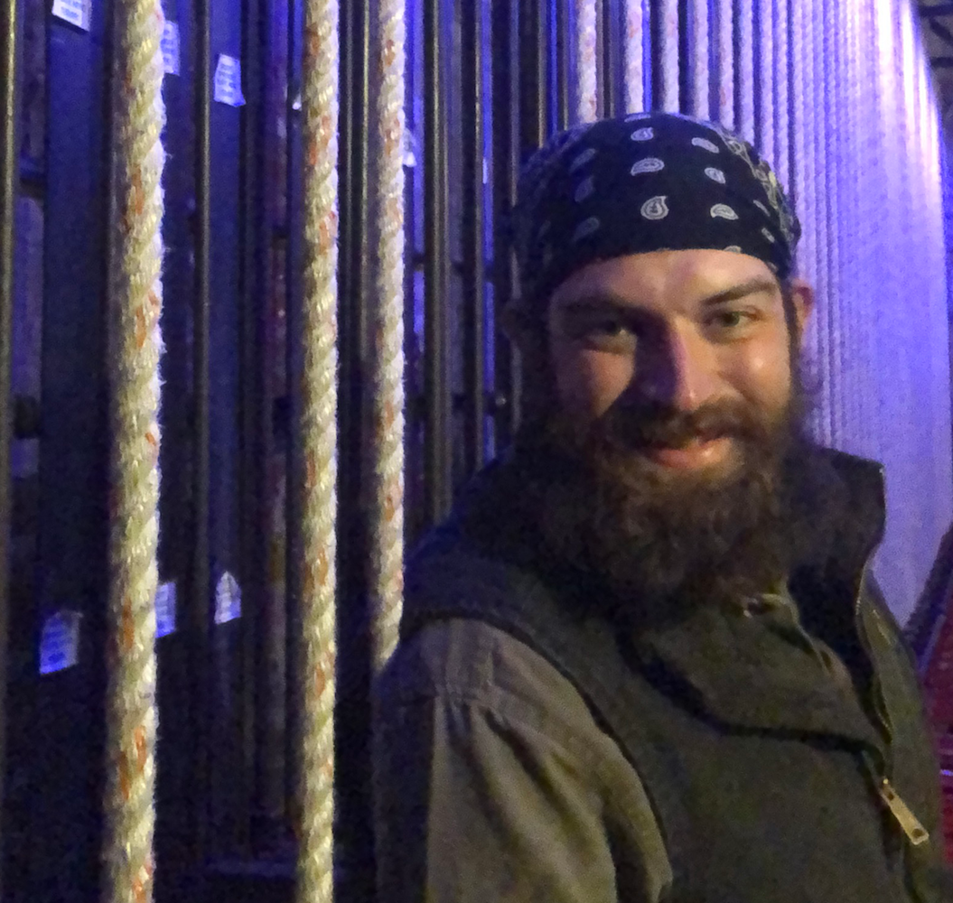 A white man with a bushy dark beard and long hair tied back under a bandana, standing at the rail and smiling towards the camera.