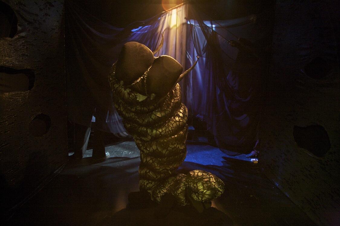 An image from Kate Brehm's show Dark Space. A human-sized, green and black chrysalis onstage, lit dramatically from above.