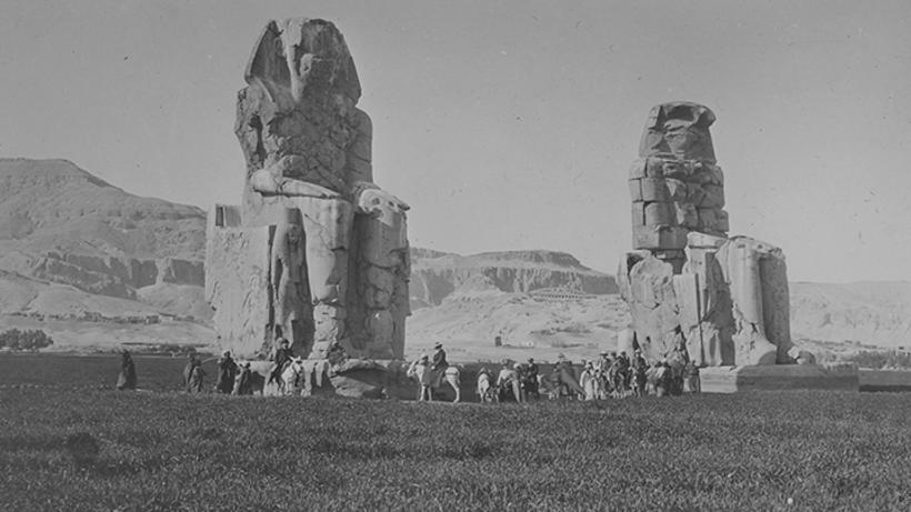 Lots of people walking or on horseback next to two massive Ancient Egyptian Statues