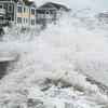 A large wave crashes against a sea wall near a row of houses.